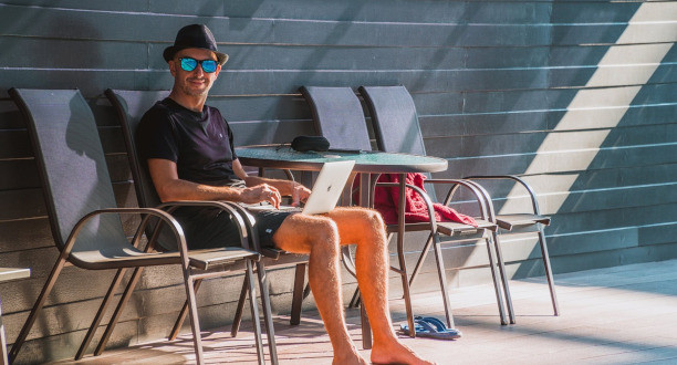 Mann sitzt mit Urlaubsoutfit auf einem Sonnenstuhl und hält Laptop auf dem Schoß.