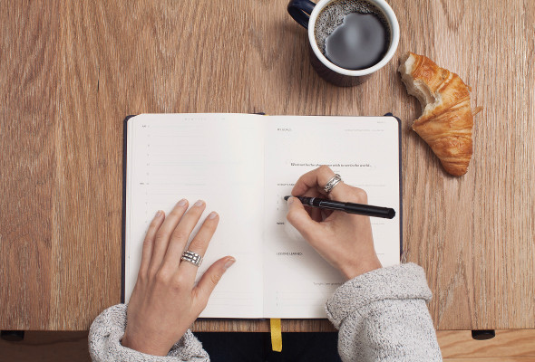 Eine Frau schreibt etwas in einen Kalender . Ein Kaffe sowie Gebäck liegt daneben. 